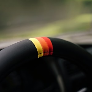 May include: Close-up of a black leather steering wheel with a red, orange, and yellow stripe detail.