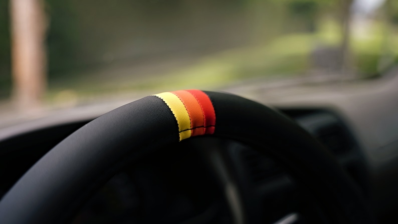 May include: Close-up of a black leather steering wheel with a red, orange, and yellow stripe detail.