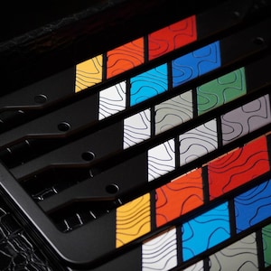 Puede incluir: Marcos de matrícula de metal negro con inserciones coloridas de estilo mapa topográfico. Las inserciones presentan una variedad de colores, incluyendo amarillo, naranja, rojo, azul, verde y plateado. Los marcos se muestran sobre una superficie negra texturizada.