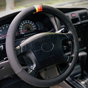 May include: Black leather steering wheel cover with a red, yellow, and black stripe design. The cover has a textured surface and is designed to fit most standard steering wheels.