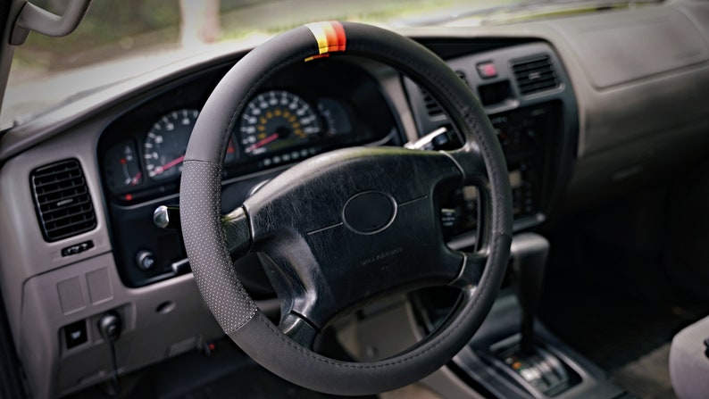 May include: Black leather steering wheel cover with a red, yellow, and black stripe design. The cover has a textured surface and is designed to fit most standard steering wheels.