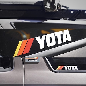 Op de afbeelding: Detail van een voertuig met een zwart paneel met het witte woord "YOTA" en een retro streepontwerp in geel, oranje en rood. Een kleinere sticker met hetzelfde ontwerp is zichtbaar op het raam.