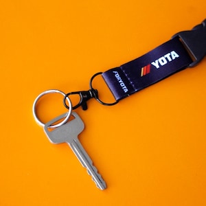 以下が含まれることがあります： 「YOTA」の文字が入った黒いストラップと鍵。ストラップには黒いプラスチック製の留め具と金属製のキーリングが付いています。鍵は銀色でシンプルなデザインです。背景は明るいオレンジ色です。