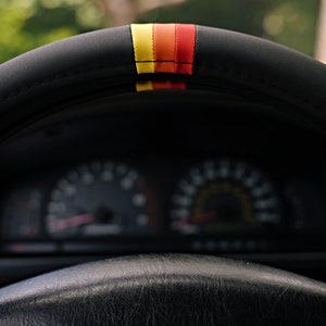 May include: A black leather steering wheel with a red, yellow, and orange stripe. The steering wheel is in a car with a dashboard visible in the background.