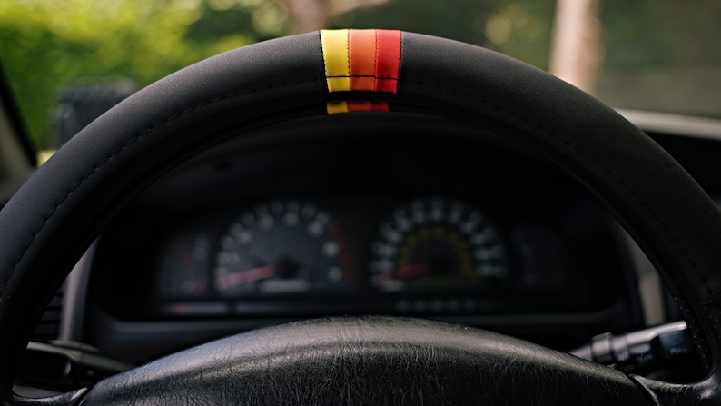 May include: A black leather steering wheel with a red, yellow, and orange stripe. The steering wheel is in a car with a dashboard visible in the background.