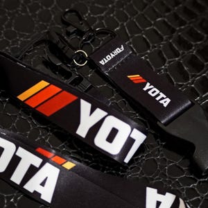 May include: Black lanyards with white "YOTA" text and orange and yellow stripes. One lanyard has a black clip and the other has a black buckle. The word "FORYOTA" is printed on the lanyard.