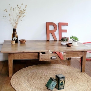 Peut inclure: Une table basse rectangulaire en bois avec un tiroir coulissant doté d'une poignée en métal. Le plateau supporte une bouteille en verre brun avec des herbes séchées, deux petits bols en bois, deux plantes succulentes et les lettres rouges R et E.