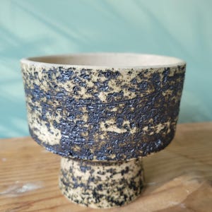 Peut inclure: Un pot en céramique avec une finition texturée et mouchetée dans des tons beige et noir. Le pot a un dessus large et rond et une base en piédestal.