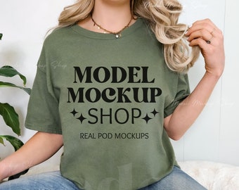 May include: A woman wearing a green t-shirt with the text "MODEL MOCKUP SHOP" and the words "REAL POD MOCKUPS" printed on it. She is sitting on a white surface wearing blue jeans.