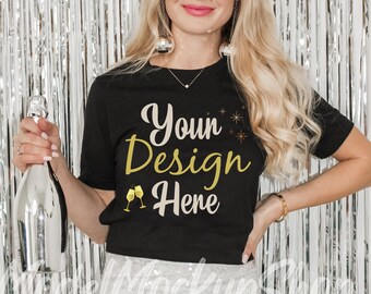 Puede incluir: Una camiseta negra con el texto "Your Design Here" en letras doradas y dos copas de champán doradas. La camiseta la lleva puesta una persona delante de un fondo de flecos plateados.
