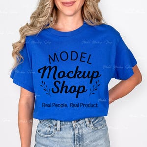 May include: A royal blue t-shirt with the words "MODEL Mockup Shop" in a black script font. Below the text is the phrase "Real People. Real Product." The model is wearing blue jeans.
