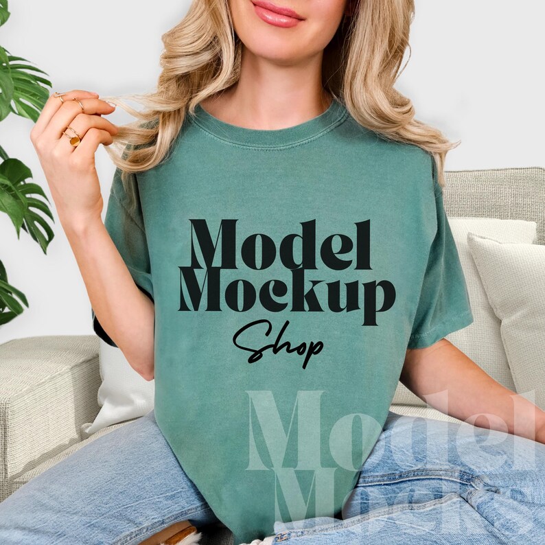 May include: A teal-colored t-shirt with the words "Model Mockup Shop" in a bold, black font. The shirt is a relaxed fit and appears to be made of soft material. The model is wearing blue jeans.