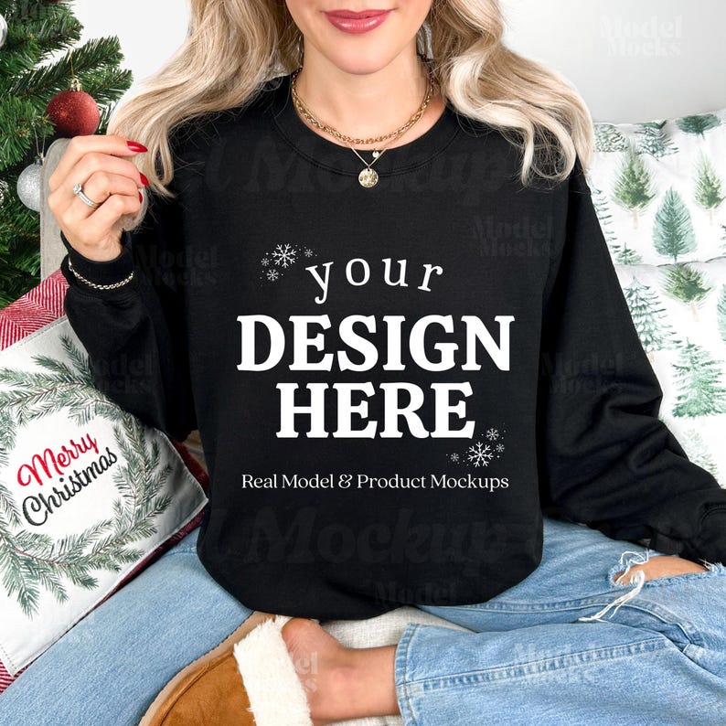 May include: A black sweatshirt with the text "your DESIGN HERE" in white, with snowflake accents. The sweatshirt is part of a product mockup, with a Christmas-themed pillow and a Christmas tree in the background.