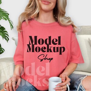 May include: Coral-colored t-shirt with the text "Model Mockup Shop" in black. The shirt is a mock-up, perfect for showcasing designs. The model is holding a white mug. The shirt is a relaxed fit.