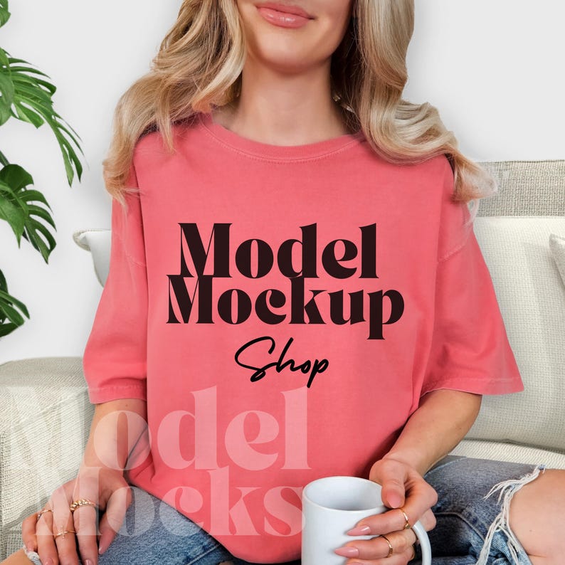 May include: Coral-colored t-shirt with the text "Model Mockup Shop" in black. The shirt is a mock-up, perfect for showcasing designs. The model is holding a white mug. The shirt is a relaxed fit.