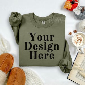 May include: Olive green sweatshirt with the text "Your Design Here" in black. The sweatshirt is displayed with fall-themed decorations, including a mug of coffee, a book, and decorative pumpkins.