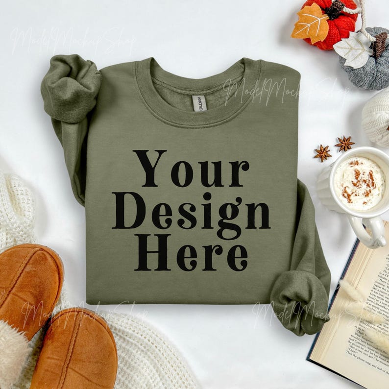 May include: Olive green sweatshirt with the text "Your Design Here" in black. The sweatshirt is displayed with fall-themed decorations, including a mug of coffee, a book, and decorative pumpkins.
