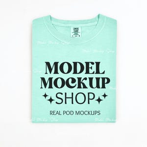 May include: A light blue t-shirt with the text "MODEL MOCKUP SHOP" in black lettering. The text is in a retro font style with two stars on either side of the word "SHOP". The text "REAL POD MOCKUPS" is printed in black below the main text.