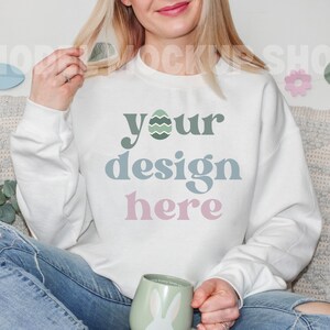 May include: White sweatshirt with the text "your design here" in green, blue, and pink. The design includes an Easter egg graphic. The person is holding a mug with a bunny design.