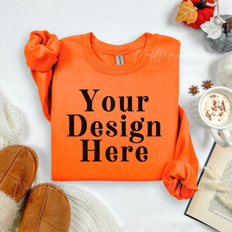 May include: Bright orange sweatshirt with the text "Your Design Here" in black. The sweatshirt is surrounded by fall-themed decorations, including a mug of coffee, a book, and decorative pumpkins.