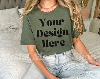 May include: A woman wearing a green short-sleeved t-shirt with the text "Your Design Here" printed on the front. She is sitting on a bed with a white bedspread.