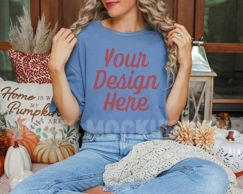 May include: A woman wearing a blue t-shirt with the text "Your Design Here" in red. She is sitting on a couch with a white pillow that says "Home is where my pumpkins are".