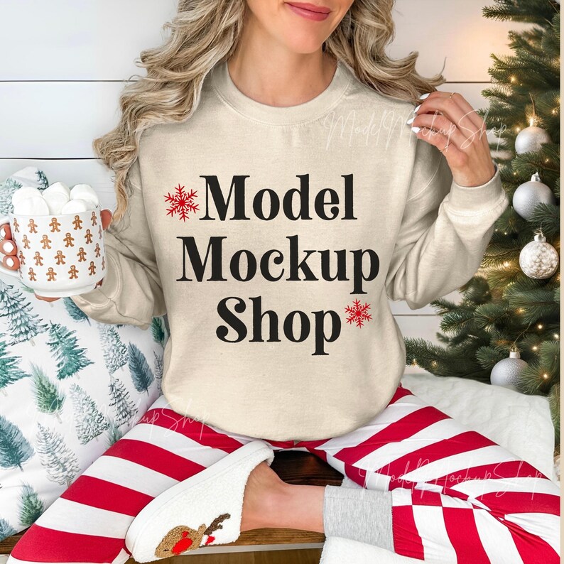 May include: A woman wearing a beige sweatshirt with the text "Model Mockup Shop" and red snowflake accents. She is wearing red and white striped pants and is sitting on a wooden surface.
