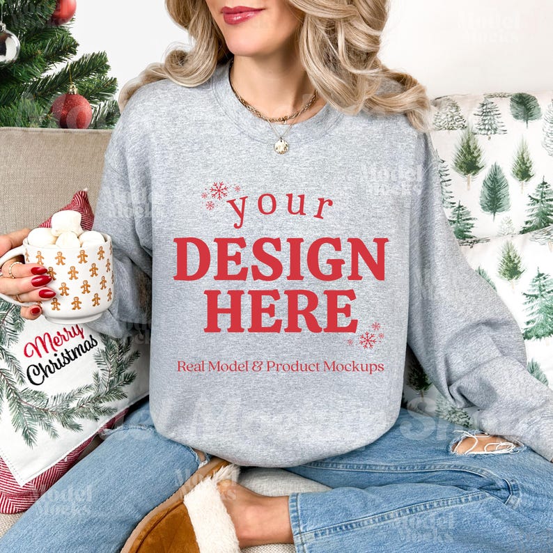 May include: A light gray sweatshirt with the text "your DESIGN HERE" in red, with snowflakes. The model is wearing blue jeans and brown slippers. A mug with gingerbread men and marshmallows is held in the hand.