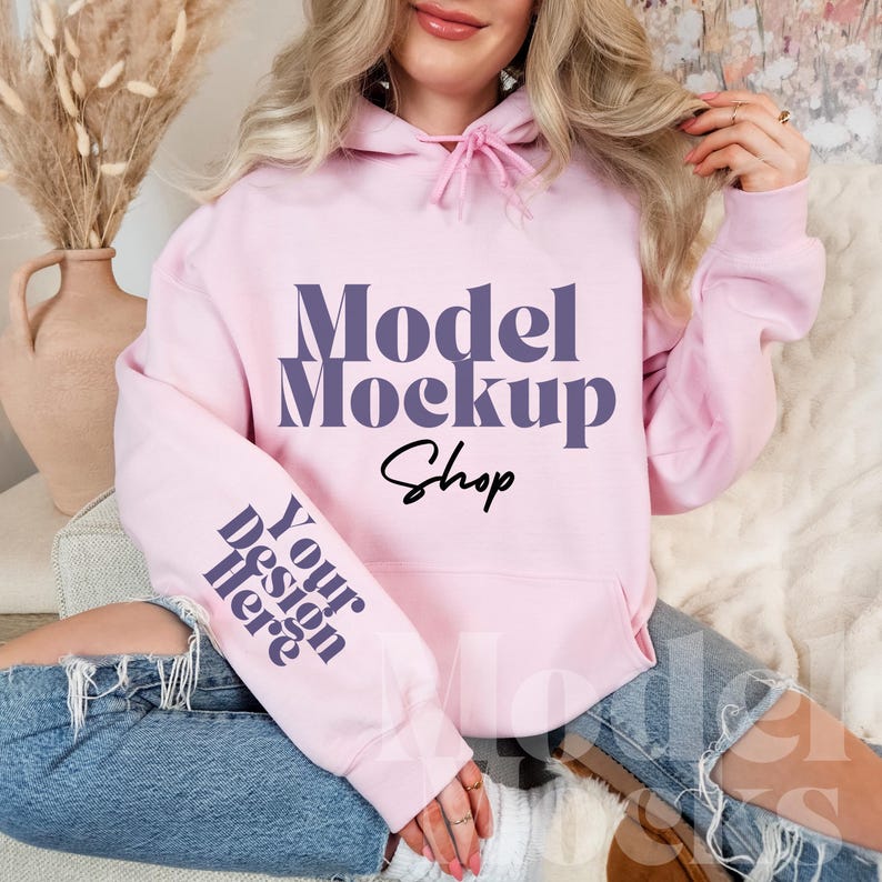 Op de afbeelding: Een roze hoodie met een paarse tekstoverlay die "Model Mockup Shop" en "Your design here" op de mouw zegt.