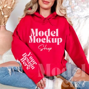 Op de afbeelding: Een rode hoodie met de tekst "Model Mockup Shop" en "Your design here" erop gedrukt. De hoodie wordt gedragen door een persoon die op een wit en bruin oppervlak zit.