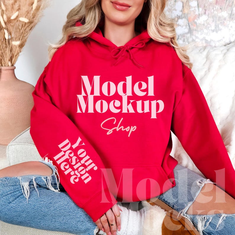 Op de afbeelding: Een rode hoodie met de tekst "Model Mockup Shop" en "Your design here" erop gedrukt. De hoodie wordt gedragen door een persoon die op een wit en bruin oppervlak zit.