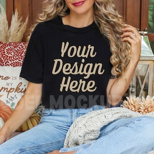 May include: A woman wearing a black t-shirt with the text "Your Design Here" in beige lettering. She is sitting on a couch with a white throw blanket and pillows.