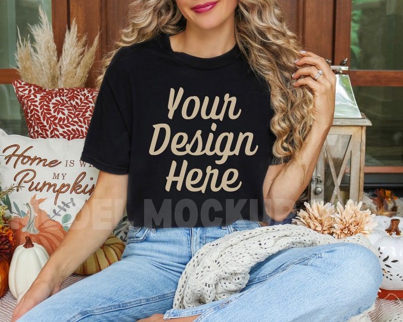 May include: A woman wearing a black t-shirt with the text "Your Design Here" in beige lettering. She is sitting on a couch with a white throw blanket and pillows.