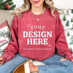 May include: A red sweatshirt with the text "your DESIGN HERE" in white. The sweatshirt is part of a product mockup, with the words "Real Model & Product Mockups" below the main text. The model is wearing blue jeans.