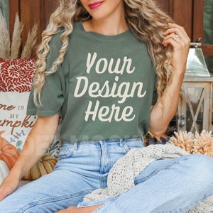 May include: A woman wearing a green t-shirt with the text "Your Design Here" in white. She is sitting on a porch with fall decorations and pumpkins.
