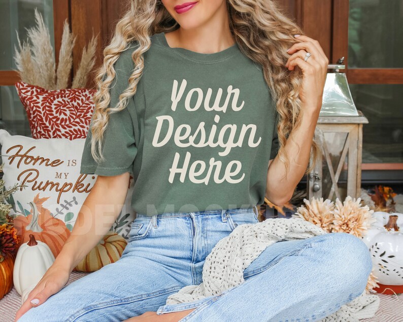 May include: A woman wearing a green t-shirt with the text "Your Design Here" in white. She is sitting on a porch with fall decorations and pumpkins.
