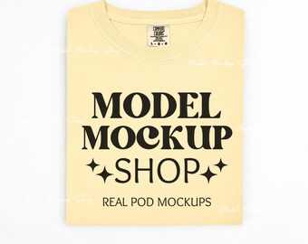 May include: A yellow t-shirt with the text "MODEL MOCKUP SHOP" in black, with two small stars on either side of the word "SHOP". The text "REAL POD MOCKUPS" is below the main text. A small tag is visible on the shirt, with the text "COMFORT COLORS" and "L-G-O".