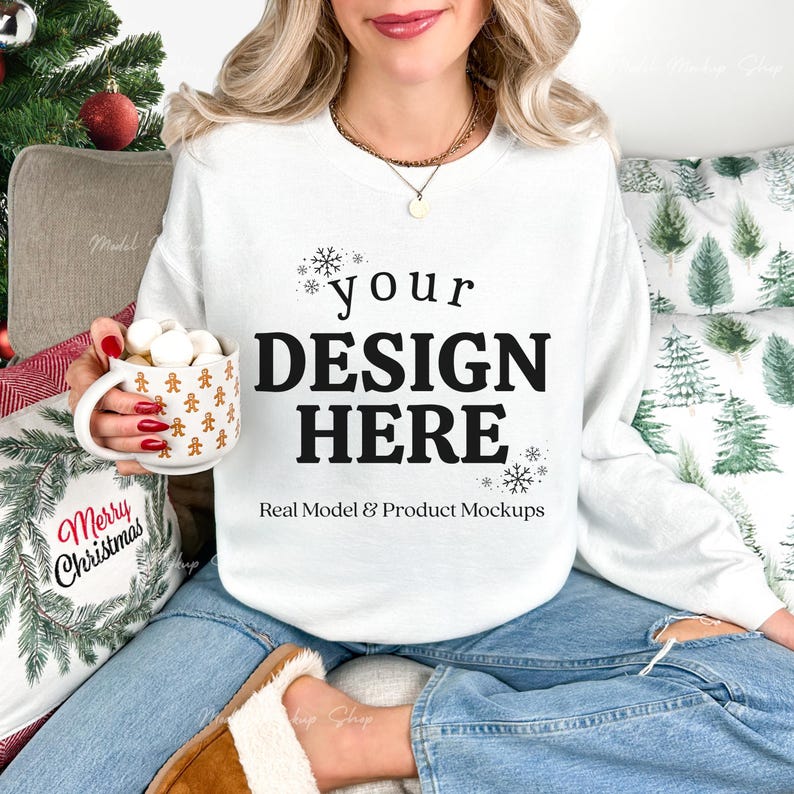 May include: White sweatshirt with the text "your DESIGN HERE" in black. The model is wearing blue jeans and brown slippers. A mug with gingerbread men is held in the hand. A pillow with "Merry Christmas" is visible.