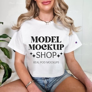 May include: A white cropped t-shirt with short sleeves featuring the text "MODEL MOCKUP SHOP" in black with a retro font and two small stars on either side of the text. The text "REAL POD MOCKUPS" is printed below the main text. The shirt is being worn by a person with long blonde hair and is sitting down.