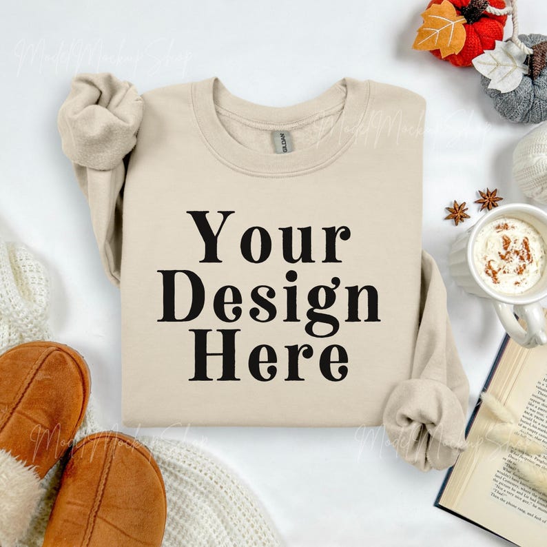 May include: A beige sweatshirt with the text "Your Design Here" in black. The sweatshirt is surrounded by fall-themed decorations, including a mug of coffee, a book, and decorative pumpkins.