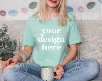 Puede incluir: Una mujer lleva una camiseta verde claro con el texto "your design here" en blanco. Está sentada en un sofá con un cojín blanco y sostiene una taza de cerámica blanca con un diseño de conejo.