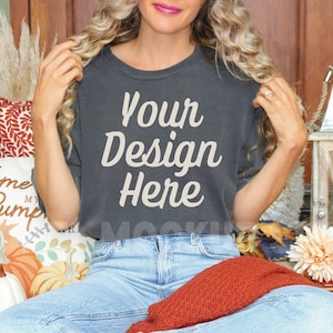 May include: A woman wearing a dark gray t-shirt with the text "Your Design Here" in beige. She is sitting on a porch with fall decor, including pumpkins and a lantern.