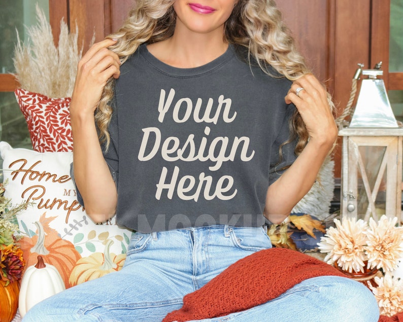 May include: A woman wearing a dark gray t-shirt with the text "Your Design Here" in beige. She is sitting on a porch with fall decor, including pumpkins and a lantern.