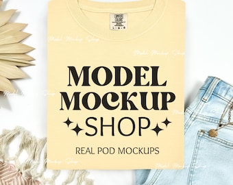 May include: A pale yellow t-shirt with the text "MODEL MOCKUP +SHOP+" and "REAL POD MOCKUPS" printed on it. The t-shirt is lying on a white surface with a pair of blue jeans and a woven straw decoration.
