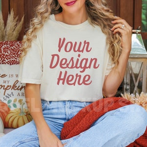 May include: A woman wearing a white t-shirt with the text "Your Design Here" in red lettering. She is sitting on a blanket with a pillow that says "Home is where my pumpkin is".