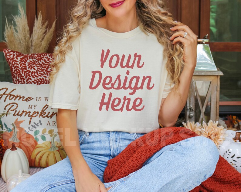 May include: A woman wearing a white t-shirt with the text "Your Design Here" in red lettering. She is sitting on a blanket with a pillow that says "Home is where my pumpkin is".