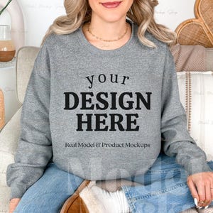 May include: A grey sweatshirt featuring the text "your DESIGN HERE" in black, bold letters. Beneath, smaller text reads "Real Model & Product Mockups". The sweatshirt is paired with blue jeans.