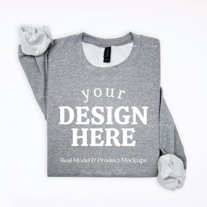 May include: A heather gray crewneck sweatshirt featuring the white text YOUR DESIGN HERE printed on the front. The garment has long sleeves with ribbed cuffs and a ribbed neckline, with a black brand label visible inside the collar area.
