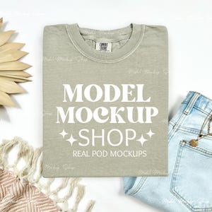 May include: A sage green t-shirt with the words "MODEL MOCKUP SHOP" in large white letters. The shirt is folded neatly on a white surface, with a pair of light blue jeans and a decorative palm leaf visible.