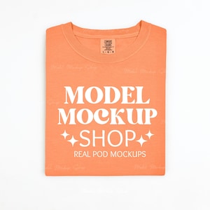 May include: A folded orange t-shirt with the text "MODEL MOCKUP SHOP" and "REAL POD MOCKUPS" printed in white. The t-shirt has a tag that says "COMFORT COLORS" and "L-G-O-G".